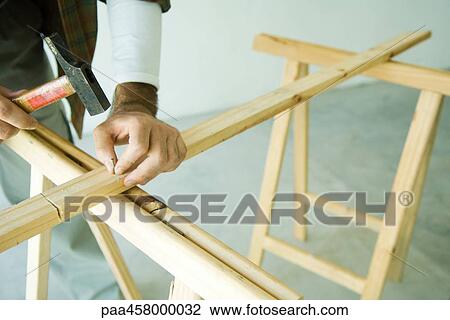 Stock Image - Man hammering nail into wood plank, cropped view. Fotosearch