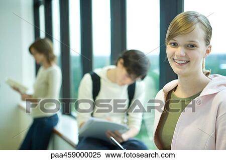 Stock Photography - Female student in front of window, smiling at camera, peers in background. Fotosearch