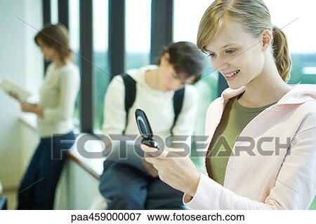 Stock Photo - Female student looking at cell phone in front of window, smiling, students in background. Fotosearch