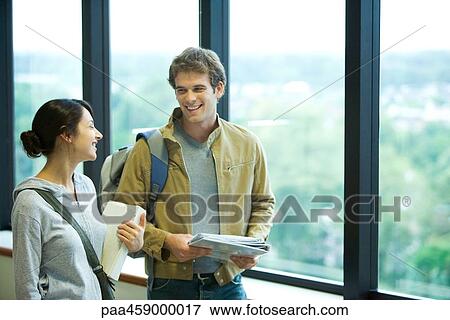 Stock Photo - Two students chatting in front of window, smiling at each other. Fotosearch