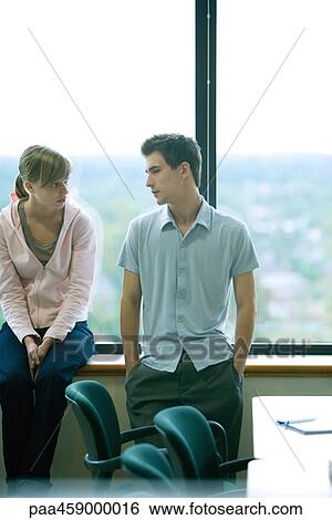 Stock Photograph - Two young friends side by side in front of window, talking, male's hand in pockets. Fotosearch