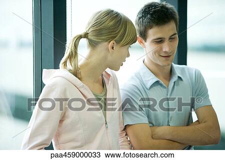 Stock Image - Two young friends standing in front of window, chatting, male's arms folded. Fotosearch