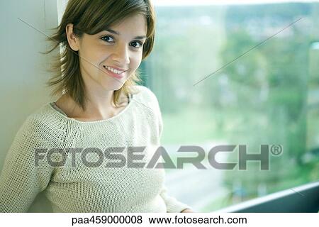 Stock Photo - Young woman sitting by window, smiling at camera. Fotosearch