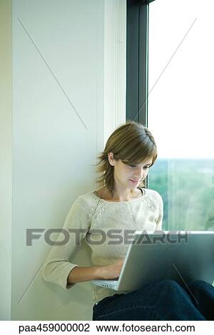 Young woman sitting by window, using laptop View Large Photo Image Stock Image - Young woman sitting by window, using laptop. Fotosearch
