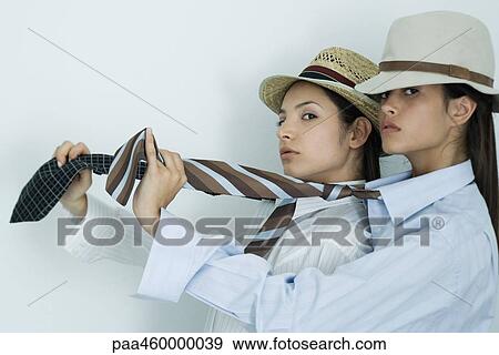 Stock Photo - Two young female friends dressed in button down shirts, ties and hats, holding out ties, glancing at camera, portrait. Fotosearch