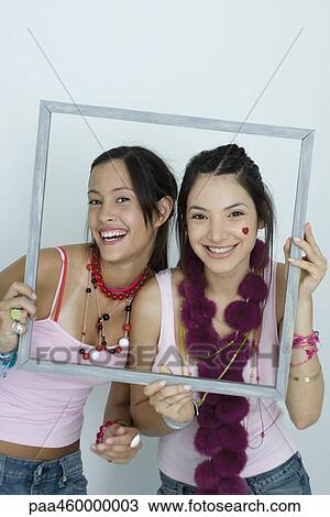 Stock Image - Two young female friends holding up picture frame, smiling at camera, portrait. Fotosearch