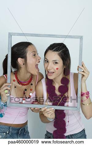Stock Photo - Two young female friends holding up picture frame, one yelling into the other's ear, portrait. Fotosearch