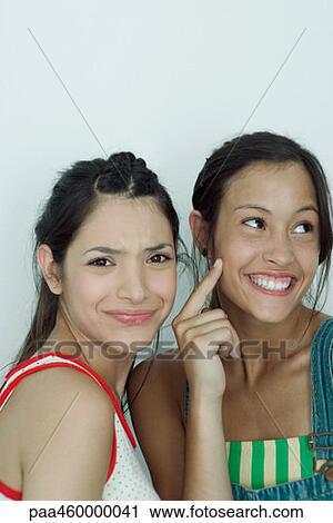 Two young female friends, one furrowing brow at camera, the other pointing to cheek, looking away, portrait View Large Photo Image Stock Image - Two young female friends, one furrowing brow at camera, the other pointing to cheek, looking away, portrait. Fotosearch