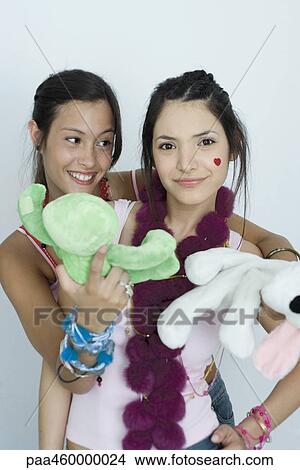 Picture - Two young female friends, one standing behind the other, holding out stuffed toys, portrait. Fotosearch