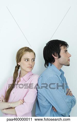 Stock Image - Young couple standing back to back with arms crossed, woman looking over shoulder, waist up, portrait. Fotosearch