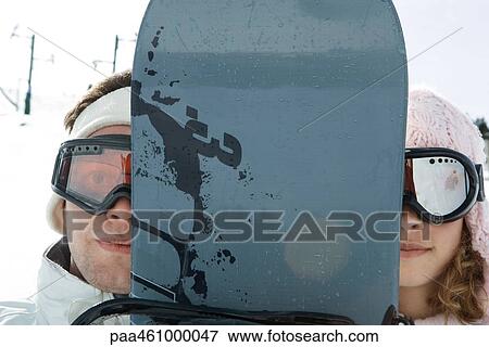 Stock Photo - Two young friends peeking at camera from behind snowboard, portrait. Fotosearch