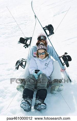 Stock Image - Two young skiers sitting under skis together, smiling at camera, portrait. Fotosearch
