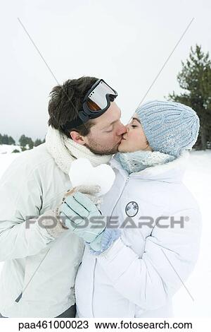Stock Image - Young couple kissing, holding heart made of snow together, eyes closed, portrait. Fotosearch