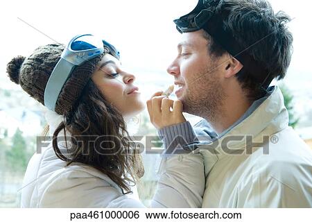 Stock Photograph - Young woman applying lipbalm to boyfriend's lips, both dressed in winter clothing, side view. Fotosearch