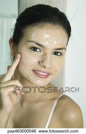 Stock Photograph - Young woman putting exfoliant mask on face, smiling at camera. Fotosearch