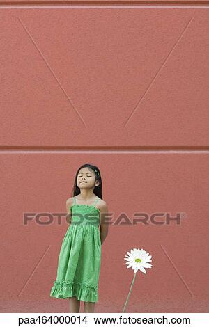 Picture - Girl standing with eyes closed and hands behind back, next to flower. Fotosearch