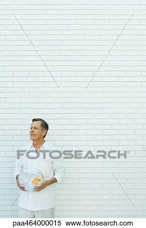 Stock Photography - Man standing with eyes closed, holding goldfish bowl, in front of white brick wall, three quarter length. Fotosearch