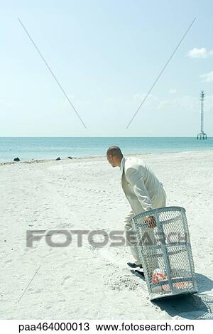 Stock Image - On beach, man in suit pulling garbage can towards ocean, side view, full length. Fotosearch