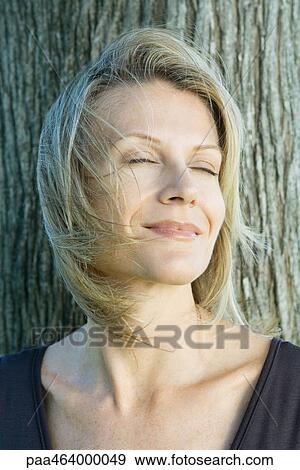 Woman smiling, eyes closed, hair blowing in wind, tree trunk in background, head and shoulders View Large Photo Image Stock Photo - Woman smiling, eyes closed, hair blowing in wind, tree trunk in background, head and shoulders. Fotosearch