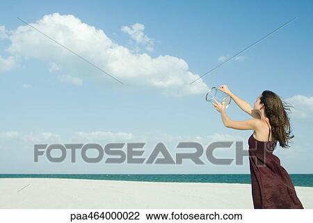 Woman standing on beach, holding up clear container, releasing clouds, optical illusion View Large Photo Image Stock Image - Woman standing on beach, holding up clear container, releasing clouds, optical illusion. Fotosearch