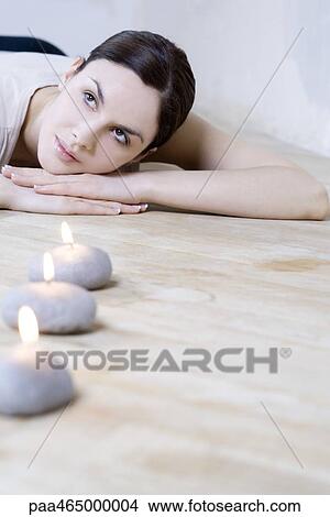 Young woman lying on stomach, head resting on arms, burning candles in foreground View Large Photo Image Picture - Young woman lying on stomach, head resting on arms, burning candles in foreground. Fotosearch