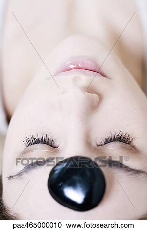 Stock Image - Young woman reclining with pebble on forehead, eyes closed, close-up. Fotosearch