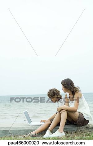 Stock Image - Boy and teen sister using laptop by water's edge. Fotosearch