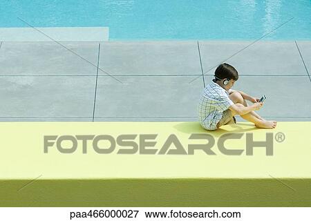 Boy sitting next to swimming pool, using handheld video device, wearing headphones View Large Photo Image Stock Photo - Boy sitting next to swimming pool, using handheld video device, wearing headphones. Fotosearch
