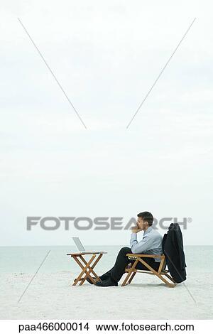Picture - Businessman sitting in chair on beach, looking at laptop computer, side view. Fotosearch