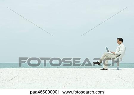 Stock Image - Businessman using laptop computer on the beach, full length. Fotosearch