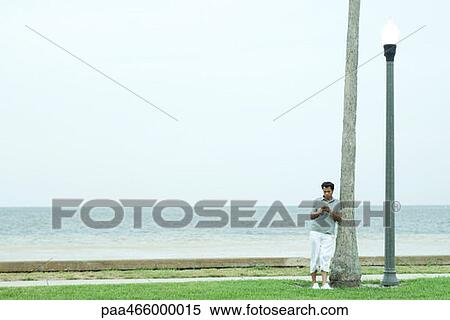 Stock Photography - Man leaning against tree trunk, looking down at cell phone, full length, mid-distance. Fotosearch