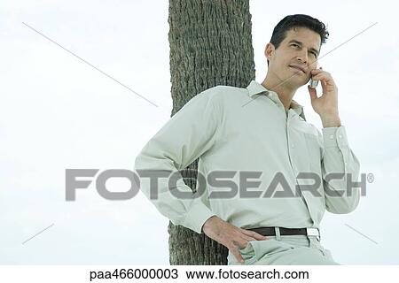 Stock Image - Man leaning against tree trunk, using cell phone, waist up. Fotosearch