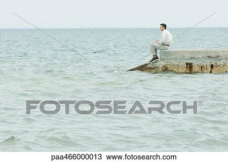 Stock Image - Man sitting on breakwater, holding laptop computer, looking at view. Fotosearch