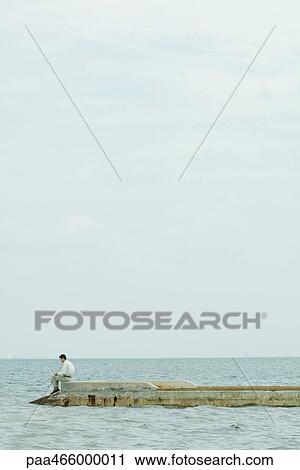 Stock Image - Man sitting on breakwater, using laptop computer, in the distance. Fotosearch