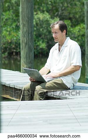 Picture - Man sitting on dock, using laptop computer, looking away. Fotosearch
