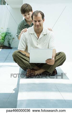 Man sitting on ground using laptop computer, son looking over his shoulder View Large Photo Image Stock Photograph - Man sitting on ground using laptop computer, son looking over his shoulder. Fotosearch