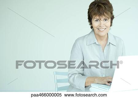 Stock Photography - Senior woman sitting, using laptop computer, smiling at camera. Fotosearch