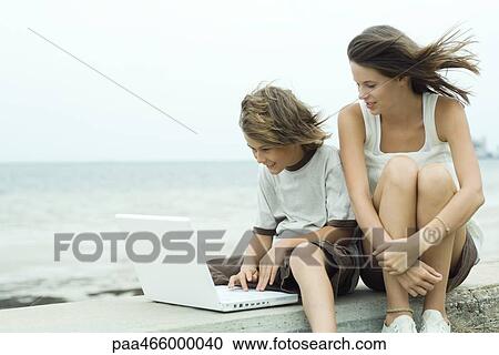 Stock Image - Teen girl and little brother using laptop near coast. Fotosearch