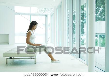 Young woman sitting on bench, using laptop computer, full length View Large Photo Image Picture - Young woman sitting on bench, using laptop computer, full length. Fotosearch