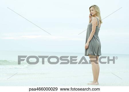 Young woman standing on beach, using cell phone, looking away View Large Photo Image Stock Photo - Young woman standing on beach, using cell phone, looking away. Fotosearch