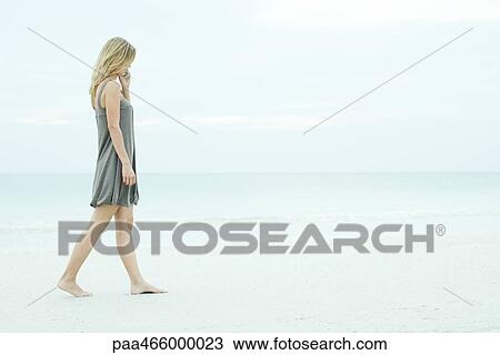 Stock Image - Young woman walking on beach, using cell phone, full length. Fotosearch