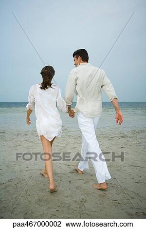 Stock Image - Father and daughter walking hand in hand on beach, rear view, full length. Fotosearch