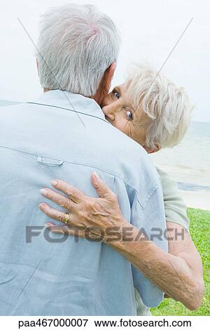 Stock Photo - Senior couple hugging, woman looking over man's shoulder at camera. Fotosearch