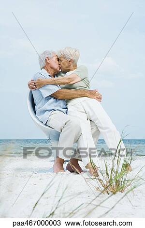 Stock Image - Senior couple sitting in chair together on beach, hugging and kissing, woman sitting on man's lap, full length. Fotosearch