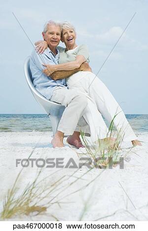 Stock Photo - Senior couple sitting in chair together on beach, embracing, woman sitting on man's lap, both smiling at camera. Fotosearch