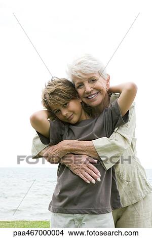 Picture - Senior woman hugging grandson from behind, both smiling at camera, portrait. Fotosearch