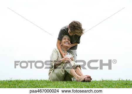 Picture - Senior woman sitting on grass, grandson standing behind her with hand on her face, giving her gift. Fotosearch