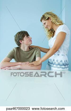 Teen boy sitting down, mother standing next to him with hand on his shoulder, smiling at each other View Large Photo Image Stock Image - Teen boy sitting down, mother standing next to him with hand on his shoulder, smiling at each other. Fotosearch