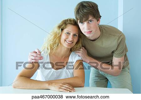Stock Photo - Teen boy standing next to mother sitting at table, cheek to cheek, smiling at camera, portrait. Fotosearch