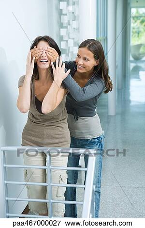 Stock Photo - Teen girl standing behind mother, hands over her eyes, both smiling, three quarter length. Fotosearch